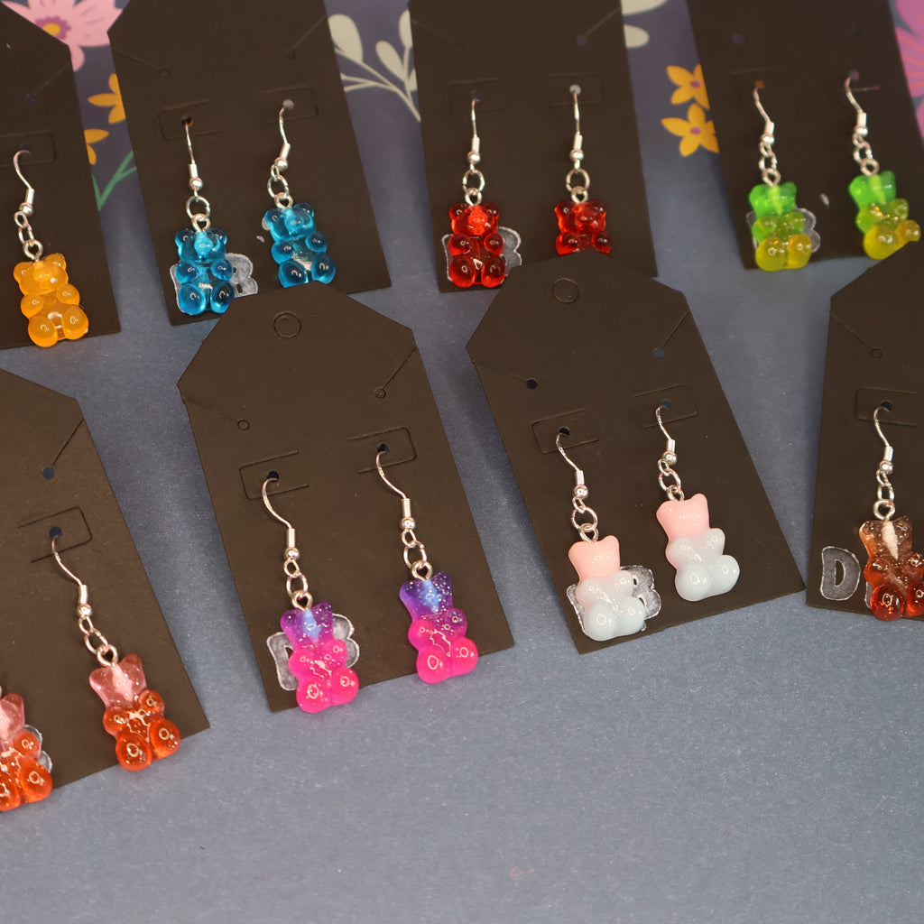 Nine pairs of dangling earrings displayed on dark brown card holders, each featuring a small translucent bear charm in different colors including orange, blue, red, green, pink, white, and brown, arranged on a gray floral surface.