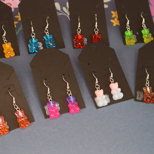 Nine pairs of dangling earrings displayed on dark brown card holders, each featuring a small translucent bear charm in different colors including orange, blue, red, green, pink, white, and brown, arranged on a gray floral surface.