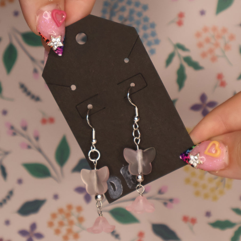 A pair of silver dangle earrings with pink butterfly charms and small pink flower charms, displayed on a black gift tag held by hands with decorated pink nails against a floral patterned background.
