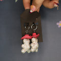 A pair of dangling earrings displayed on a black card, held by a hand with decorated nails against a dark gray floral background. The earrings feature two small glittery teddy bear charms wearing pink cowboy hats.