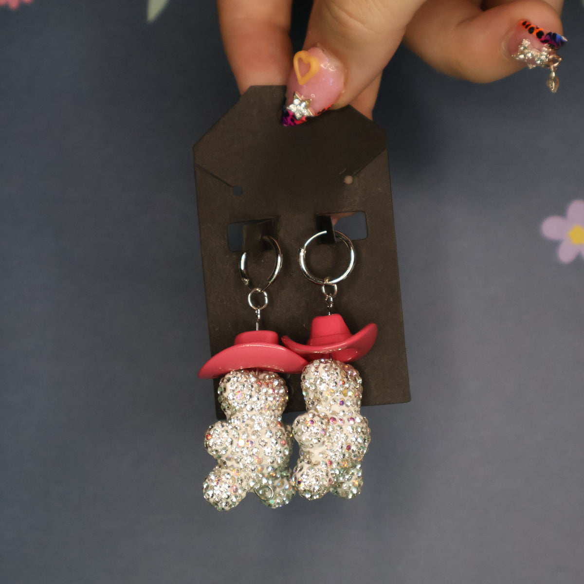 A pair of dangling earrings displayed on a black card, held by a hand with decorated nails against a dark gray floral background. The earrings feature two small glittery teddy bear charms wearing pink cowboy hats.